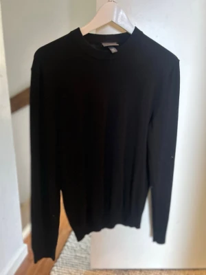 Svart stickad tröja med rund hals - En stilren svart stickad tröja med rund hals och långa ärmar. Tröjan har en klassisk passform och ribbade muddar vid ärmslut och nederkant. Perfekt att matcha med jeans eller kjol för en clean look.
