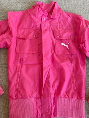 Rosa vindjacka från Puma - Snygg rosa vindjacka från Puma med vita loggan på bröstet. Jackan har två bröstfickor med knappar, två sidofickor med dragkedja och ribbade muddar vid ärmslut och nederkant. Perfekt för dig som vill sticka ut med färg.