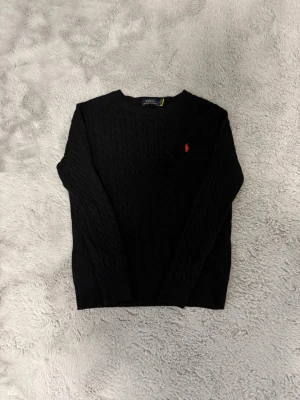Svart kabelstickad tröja Polo Ralph Lauren - Svart kabelstickad tröja från Polo Ralph Lauren med rund halsringning och röd broderad logga på bröstet. Tröjan har långa ärmar och klassisk passform, perfekt för en stilren look.