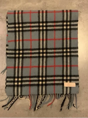Burberry halsduk - Klassisk halsduk i mjuk ull från Burberry med blå bas och rutigt mönster i svart, vitt och rött. Halsduken har fransar i ändarna och en diskret Burberry-etikett. Perfekt accessoar för att lyfta din outfit.