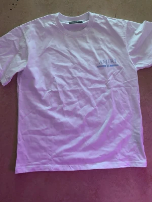 Vit AMIRI t-shirt med blå logga - Snygg vit t-shirt från AMIRI med blå logga tryckt på bröstet och större tryck på ryggen. Klassisk passform med rund hals och korta ärmar. Perfekt för dig som gillar stilrena och trendiga plagg med exklusiv känsla.