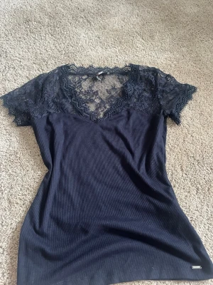 Mörkblå topp med spetsdetaljer - Snygg marinblå topp med ribbad struktur och v-ringning. Överdelen och ärmarna är i genomskinlig spets med blommönster, vilket ger en elegant touch. Perfekt att styla till jeans eller kjol för en trendig look.