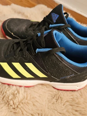 Svarta Adidas sneakers med blå detaljer - Snygga svarta sneakers från Adidas med klassiska gula ränder på sidorna, blå insida och röda detaljer på sulan. Skorna har snörning och perforerade partier för extra ventilation. Perfekta för dig som gillar sportig stil och vill sticka ut lite extra.