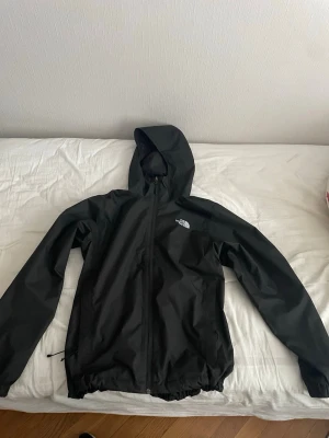 Svart regnjacka från The North Face - Snygg svart regnjacka från The North Face med huva och dragkedja framtill. Jackan har elastisk nederkant och logga på bröstet samt ryggen. Tillverkad i vattenavvisande material, perfekt för regniga dagar.