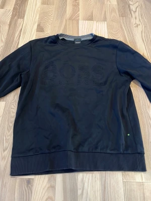 Svart sweatshirt från Hugo boss - Svart sweatshirt från Hugo boss med diskret logotyp i svart på bröstet. Tröjan har rund halsringning, ribbade muddar och är tillverkad i mjukt material. Perfekt för en stilren och avslappnad look.