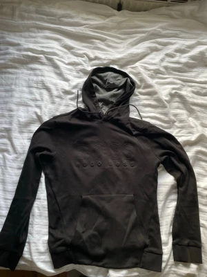 Svart hoodie från Hugo Boss - Säljer en svart hoodie från Hugo Boss med stor BOSS-logga broderad på bröstet. Hoodien har huva med dragsko, känguruficka framtill och ribbade muddar vid ärmar. I storlek M