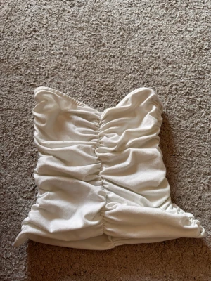 Vit bandeau tubtopp med rynk - Snygg vit bandeau tubtopp med rynkade detaljer framtill och baktill. Toppen är axelbandslös och har en tight passform som framhäver siluetten. Perfekt att styla med jeans eller kjol för en trendig look.