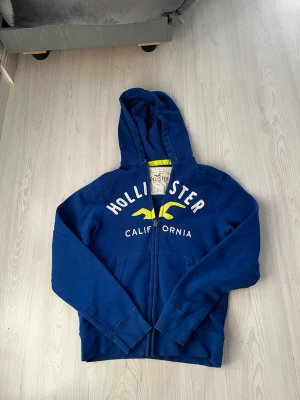 Blå Hollister hoodie med dragkedja - Snygg blå hoodie från Hollister med vit och gul logga på bröstet och texten 'California'. Dragkedja framtill, två fickor och huva. Perfekt för en avslappnad och cool stil. Mjuk bomullskänsla och klassisk passform. Inga snören tyvärr, Storlek L men passar på Medium också