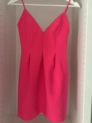 Rosa kort klänning med smala axelband - Snygg rosa kort klänning med smala axelband och v-ringning. Klänningen har en enkel, stilren design och är figurnära med lätt markerad midja. Perfekt för dig som vill sticka ut med en färgstark look. Klänningen är tyvärr uppsydd i axelbanden vilket gör att det blir lite tajt i armarna
