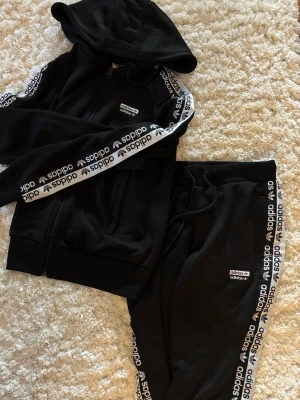 Svart Adidas mjukis-set - Svart set från Adidas med matchande hoodie och byxor. Båda plaggen har vita Adidas-loggor och text längs ärmar och ben. Hoodien har dragkedja, byxorna har elastisk midja med snörning. Mjukt och bekvämt material