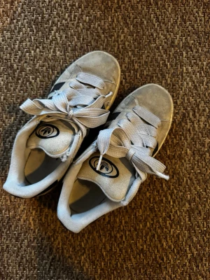 Grå Adidas sneakers med svarta ränder - Snygga Adidas sneakers i grå mocka med klassiska svarta ränder på sidorna och breda vita skosnören. Låg modell med vit sula och bekväm passform. Perfekta för en avslappnad streetstyle-look.