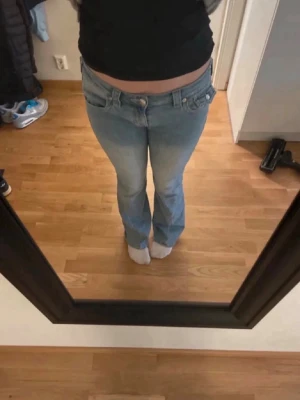Ljusblå bootcut jeans från true religion  - Säljer ett par ljusblå bootcut jeans från true religion med låg midja och stora fickor bak med knappar. Jeansen har klassisk femficksdesign och är tillverkade i denim med lätt tvättad look. Perfekta för dig som gillar retrostil och vill ha ett par snygga jeans med detaljer.
