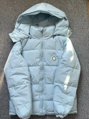 Ljusblå pufferjacka från Trapstar - Snygg ljusblå pufferjacka från Trapstar med huva och dragkedja framtill. Jackan har ett broderat Trapstar-tryck på ryggen och en rund logga på bröstet. Perfekt för kalla vinterdagar och ger en cool streetwear-look.