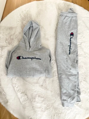 Grått Champion loungewear-set - Mysigt grått loungewear-set från Champion med matchande hoodie och byxor. Hoodien har logga framtill och byxorna har logga längs benet. Perfekt för chill dagar hemma. Mjuk bomullsblandning och avslappnad passform.