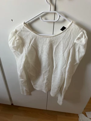 Vit långärmad blus med knyt - Stilren vit blus från Lindex med lång ärm och lätt rynkad struktur. Blusen har en rundad halsringning med knytband framtill och är tillverkad i ett tunt, mjukt tyg som ger en avslappnad men chic look. Perfekt att matcha med jeans eller kjol.