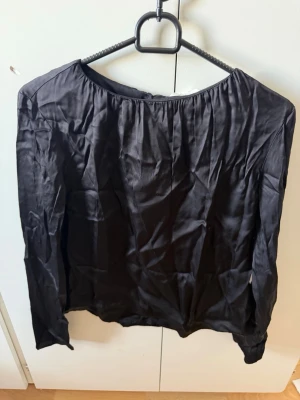 Svart långärmad blus från Object - Stilren svart blus från Object med rund halsringning och knäppning bak i nacken. Tillverkad i ett glansigt, tunt material som ger en elegant känsla. Perfekt att matcha med jeans eller kjol för en trendig look.