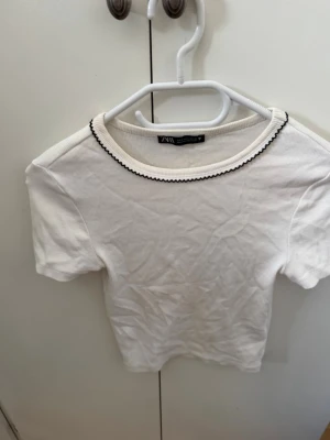 Vit ribbad topp från Zara - Säljer en vit ribbad topp från Zara med korta ärmar och en söt svart vågig kant runt halsringningen. Toppen är i mjuk bomull och har en slim passform som sitter snyggt på kroppen. Perfekt till jeans eller kjol!