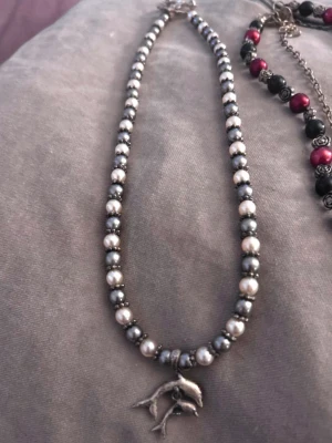Pärlhalsband med delfinberlock - Snyggt halsband med växlande vita och mörkgrå pärlor samt en silverfärgad delfinberlock. Halsbandet har en klassisk pärlform och ger en cool, havsinspirerad vibe. Perfekt accessoar för att lyfta din stil.