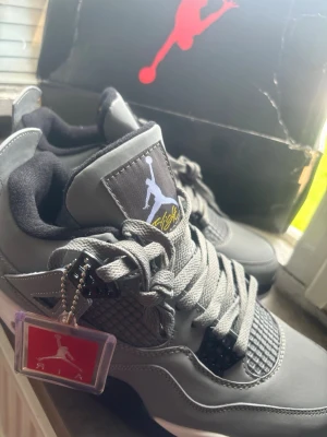 Gråa Air Jordan 4 Retro sneakers - Säljer ett par gråa Air Jordan 4 Retro sneakers med klassisk Jumpman-logga på plösen och tag. Skorna har meshdetaljer, svarta och vita inslag samt snörning. Perfekta för dig som vill ha en ikonisk och stilren sneaker i garderoben.