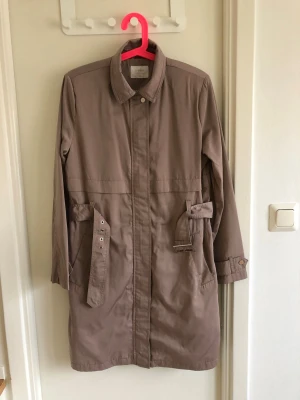 Beige trenchcoat med bälte - Stilren beige trenchcoat med klassisk krage och knäppning framtill. Jackan har bälte i midjan och två fickor med lock. Lång modell med justerbara ärmslut. Perfekt för lager-på-lager och snygg till alla outfits.