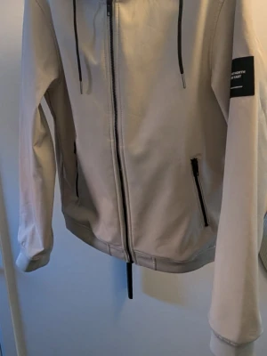 Beige vindjacka med huva - Stilren beige vindjacka med dragkedja framtill, två fickor med svarta dragkedjor och justerbar huva med snören. Jackan har en patch på ärmen och är tillverkad i ett slätt, vindtätt material. Perfekt för blåsiga dagar.