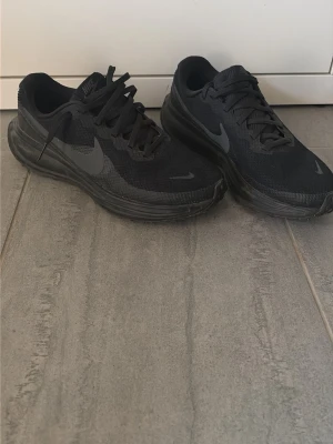 Svarta Nike sneakers med mesh - Säljer ett par helsvarta Nike sneakers med klassisk swoosh-logga på sidan. Skorna har lågt skaft, ovandel i mesh och syntetmaterial samt grov sula för bra grepp. Perfekta för en sportig och stilren look.