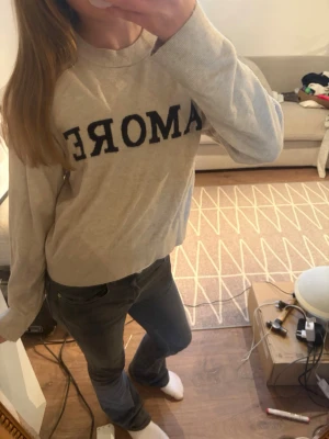 Beige stickad tröja med text - Beige stickad tröja med svart texten 'AMORE' framtill. Tröjan har rund halsringning, långa ärmar och ribbade muddar vid ärmslut och nederkant. Perfekt för en avslappnad och trendig look.