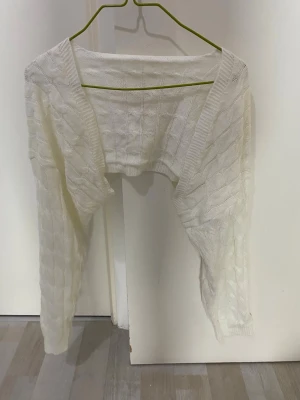 Vit kabelstickad bolero - Säljer en vit kabelstickad bolero med lång ärm och öppen front. Den är kort i modellen och har ett klassiskt stickat mönster. Perfekt att slänga över en topp för en extra touch. Materialet känns mjukt och lätt.