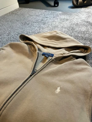 Beige hoodie från Polo Ralph Lauren - Snygg beige hoodie från Polo Ralph Lauren med dragkedja och klassisk huva. Tröjan har två fickor framtill och den ikoniska vita logon broderad på bröstet. Perfekt för en avslappnad och stilren look. Tillverkad i mjukt bomullsmaterial.