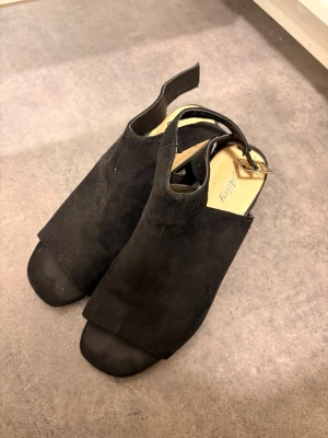 Svarta sandaler med klack från Nelly - Snygga svarta sandaler från Nelly med öppen tå och bred klack. Skorna har en stilren design i mockaimitation och justerbar rem med spänne runt hälen. Perfekta för dig som vill ha en trendig och bekväm look till sommaren.