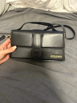 Svart Jacquemus handväska i läder - Säljer en svart handväska från Jacquemus i slätt läder med guldfärgad logga på framsidan. Väskan har ett stilrent, rektangulärt format med både handtag och axelrem. Stängs med ett lock och har en praktisk ficka på baksidan.
