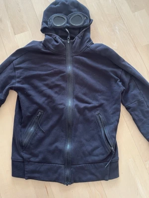Svart hoodie med glasögon i huvan - Cool svart hoodie från C.P. Company med hel dragkedja, två snedställda fickor med dragkedja och unik huva med inbyggda glasögon. Tillverkad i mjuk bomullsblandning. Perfekt för dig som gillar streetwear och vill sticka ut.