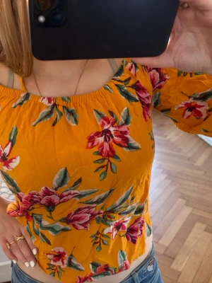 Orange offshoulder topp - Superfin gul offshoulder topp från C&A med blommigt mönster i rött, grönt och vitt. Toppen har tre kvarts ärmar och är croppad, perfekt för en färgglad och somrig look. Materialet känns lätt och luftigt.