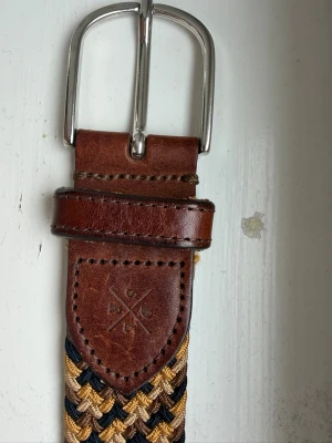 Flätat bälte från Saddler i skinn - Snyggt flätat bälte från Saddler i brunt skinn med detaljer i mörkblått, beige och gult. Spännet är i silverfärgad metall och bältet har en klassisk design som passar till många olika outfits.