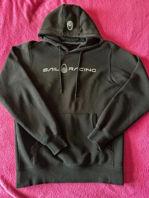 Svart hoodie från Sail Racing - Svart hoodie från Sail Racing med vit logotyp på bröstet och huvan. Klassisk design med huva, dragsnören och stor magficka. Mjuk och skön bomullsblandning som passar perfekt till chill eller streetwear. Ribbstickade muddar vid ärmslut och nederkant.