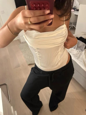 Vit satin korsett/body med tunna axelband - Snygg vit korsett-body i satin med tunna axelband och draperad urringning. Figurnära passform som ger en trendig siluett. Helt oanvänt med prislapp kvar, i storlek S.