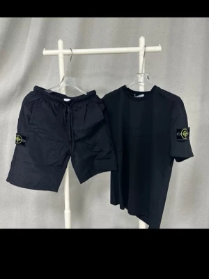 Svart set från Stone Island - Snyggt svart set från Stone Island med t-shirt och shorts. Båda plaggen har den ikoniska Stone Island-loggan på ärm och ben. T-shirten är enkel och stilren, shortsen har dragsko i midjan. Perfekt för en avslappnad och trendig look.