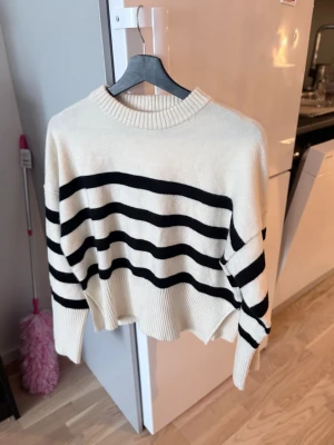 Vit stickad tröja med svarta ränder - Säljer en vit stickad tröja med breda svarta horisontella ränder. Tröjan har en avslappnad passform, ribbad rund halsringning och ribbade muddar. Perfekt för dig som gillar en klassisk och trendig look. Materialet känns mjukt och bekvämt.
