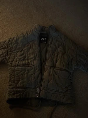 Grå quiltad jacka från Zara - Quiltad grå jacka från Zara med vågigt mönster, dragkedja framtill och stora fickor. Jackan har en boxig passform och står ut med sin trendiga, vadderade look. Perfekt för lager-på-lager-stil under kyliga dagar. (Pris kan diskuteras💕)