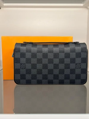 Grå/Svart handväska - Snygg handväska i rutig mönster. Väskan är svart och grå med klassiskt rutigt tryck, tillverkad i canvas med detaljer i svart läder och silverfärgad dragkedja. Perfekt accessoar för en trendig look.