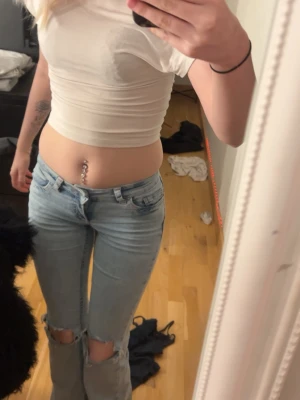 Ljusblå bootcut jeans med slitningar - Säljer ett par ljusblå bootcut jeans med slitna knän och klassisk femficksdesign. Jeansen har en röd broderad detalj på bakfickan och är i ett mjukt denimtyg. Perfekta för en avslappnad och trendig look.