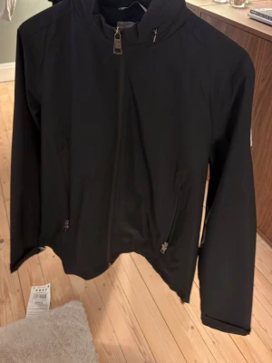 Svart vindjacka från Moncler - Snygg svart vindjacka från med dragkedja framtill och två fickor med dragkedja. Har även en luva som går att ta ut i bak. Jackan har en stilren design och logga på ärmen. NFC tagg finns och även label inne i jackan!