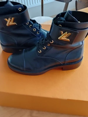 Svarta Louis Vuitton kängor Flat ranger - Snygga svarta kängor från Louis Vuitton  med guldfärgade LV-detaljer på skaftet. Klassisk snörning och robust sula gör dem både stilrena och trendiga. Perfekta för dig som vill ha en exklusiv look med streetkänsla.