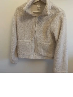 Beige teddyjacka med dragkedja - Mysig beige teddyjacka med hög krage och dragkedja framtill. Jackan har två stora fickor på framsidan och är tillverkad i fluffigt teddy-material som håller dig varm. Perfekt för kyliga dagar och enkel att matcha med olika outfits.
