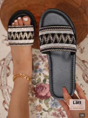 Svarta sandaler med broderad rem - Svarta sandaler med bred broderad rem i svart, vitt och guld. Snyggt mönster med zickzack-detaljer och bekväm platt sula. Perfekta för sommaren och lätta att matcha med olika outfits.