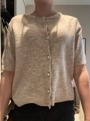Beige kofta med pärlknappar - Kortärmad beige kofta med rund halsringning och pärlknappar framtill. Mjuk och lättstickad, perfekt för lager-på-lager. Enkel och stilren design som passar till många outfits.❤️🌺