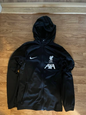 Svart Nike Liverpool hoodie - Storlek 147-159. Svart hoodie med dragkedja från Nike, officiell Liverpool FC-träningströja med LFC-logga och AXA-sponsortryck på bröstet. Tillverkad i ett glansigt, sportigt material och har två fickor framtill samt huva.