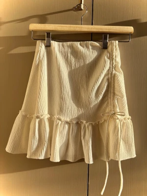 Krämvit volangkjol med knyt - Supersöt kort kjol i krämvitt med volangkant och knyt i sidan. Kjolen har en lätt textur i tyget. Perfekt att styla med en enkel topp eller stickad tröja.