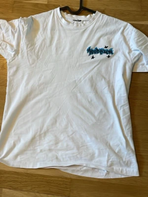 Vit Hoodrich t-shirt med tryck - Vit t-shirt från Hoodrich med flera coola tryck i blått och svart både fram och bak. T-shirten har klassisk passform och rund halsringning. Perfekt för dig som gillar streetwear och grafiska detaljer.