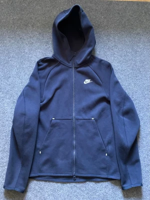 Nike tech mörkblå - perfekt skick - stl XS (passar även till Small) - Snygg mörkblå hoodie från Nike med hel dragkedja och klassisk vit logga på bröstet. Tröjan har huva och två stora fickor framtill. Perfekt för en sportig och avslappnad stil. Tillverkad i mjukt och bekvämt material.